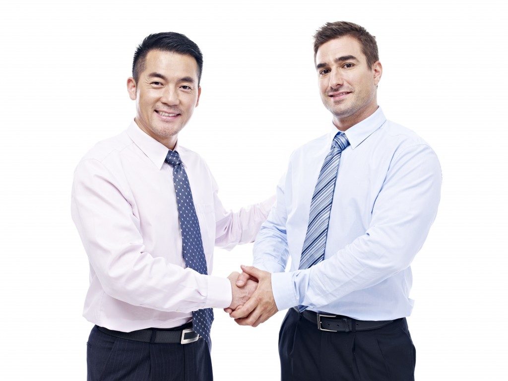asian and caucasian businessmen shaking hands looking at camera smiling, isolated on white background.